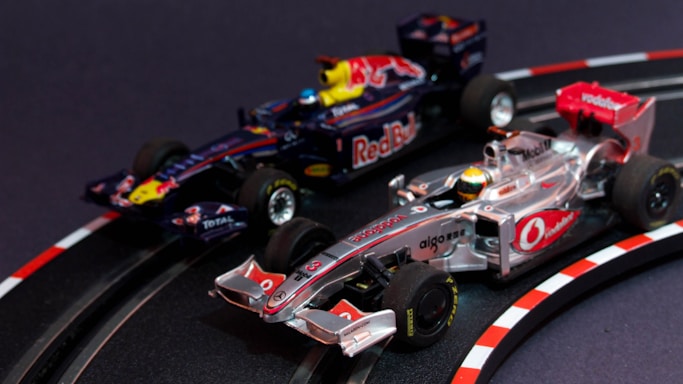 Two model Formula 1 cars are seen racing on a miniature track. The cars are intricately detailed, with one being silver and red, while the other is dark blue with red bull branding. The track features realistic racing elements such as a curve with guardrails and directional markings.
