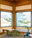 A serene indoor dining area featuring large windows with a view of lush greenery and mountains. The room is adorned with wooden frames and furniture including a table surrounded by four chairs. There are potted plants placed strategically, adding a touch of nature to the ambiance.