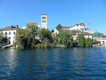 A picturesque lakeside scene featuring historic buildings with stone and plaster facades, lush green trees, and a prominent tower. The water is calm and reflects the blue sky above.