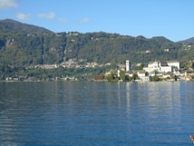 A picturesque lakeside town bordered by lush green hills. The town features charming, historical buildings, including a prominent structure with a tower. The reflection of these buildings shimmers on the tranquil water of the lake.