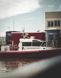 A pilot boat with the name 'Itajai & Navegantes Pilots' is docked near a waterway. The boat is white with red detailing and has two lifebuoys visible on its side. In the background, there are industrial buildings, a container, and a large blue water tank with the word 'FORTLEV' written on it. The sky is cloudy with some patches of blue.
