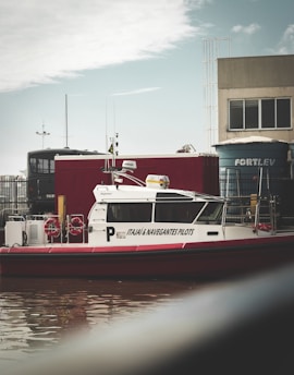 A pilot boat with the name 'Itajai & Navegantes Pilots' is docked near a waterway. The boat is white with red detailing and has two lifebuoys visible on its side. In the background, there are industrial buildings, a container, and a large blue water tank with the word 'FORTLEV' written on it. The sky is cloudy with some patches of blue.