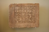 An ancient tablet with a grid of carved symbols or characters in an unknown script, mounted on a pale wall using four white brackets.