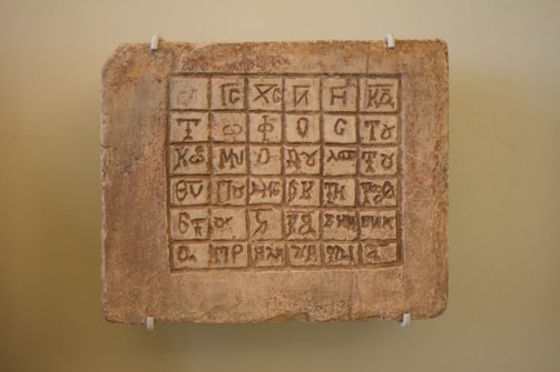An ancient tablet with a grid of carved symbols or characters in an unknown script, mounted on a pale wall using four white brackets.