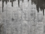A textured concrete wall with dark water stains creating irregular patterns that resemble icicles or stalactites. The wall's surface features various shades of gray with a rough, aged appearance.