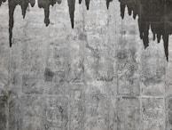 A textured concrete wall with dark water stains creating irregular patterns that resemble icicles or stalactites. The wall's surface features various shades of gray with a rough, aged appearance.