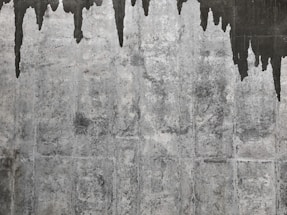A textured concrete wall with dark water stains creating irregular patterns that resemble icicles or stalactites. The wall's surface features various shades of gray with a rough, aged appearance.