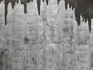 A textured concrete wall with dark water stains creating irregular patterns that resemble icicles or stalactites. The wall's surface features various shades of gray with a rough, aged appearance.