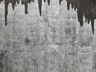 A textured concrete wall with dark water stains creating irregular patterns that resemble icicles or stalactites. The wall's surface features various shades of gray with a rough, aged appearance.