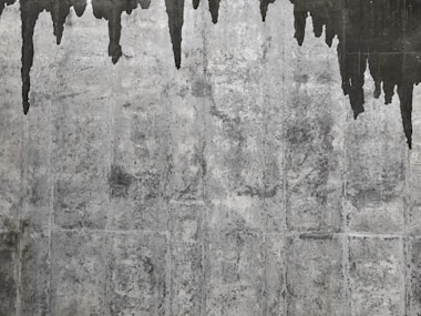 A textured concrete wall with dark water stains creating irregular patterns that resemble icicles or stalactites. The wall's surface features various shades of gray with a rough, aged appearance.