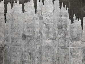 A textured concrete wall with dark water stains creating irregular patterns that resemble icicles or stalactites. The wall's surface features various shades of gray with a rough, aged appearance.