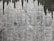 A textured concrete wall with dark water stains creating irregular patterns that resemble icicles or stalactites. The wall's surface features various shades of gray with a rough, aged appearance.
