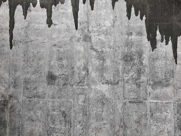 A textured concrete wall with dark water stains creating irregular patterns that resemble icicles or stalactites. The wall's surface features various shades of gray with a rough, aged appearance.