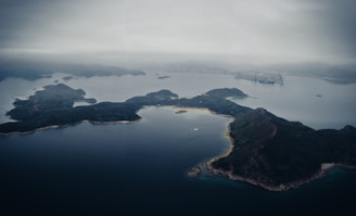 Aerial view of a series of islands covered in dense, dark green vegetation surrounded by calm, dark blue waters. The overcast sky casts a gray hue, creating a moody atmosphere. In the distance, a hint of an industrial area with structures and cranes is visible, possibly part of a port.