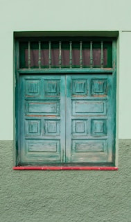 A weathered green wooden door with a rectangular window at the top, featuring vertical bars. The door is set within a green plastered wall, with a red trim at the base.