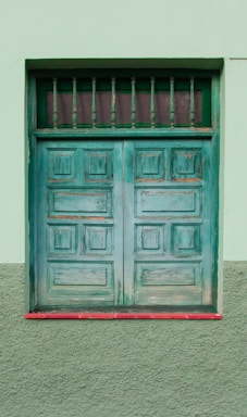 A weathered green wooden door with a rectangular window at the top, featuring vertical bars. The door is set within a green plastered wall, with a red trim at the base.