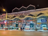 A lively street market scene at night featuring a building with the sign 'CHỢ SÔNG BẰNG'. There are various food stalls lit with bright lights, a few parked motorcycles, and people engaging in activities. Neon lights and trees are visible along the sidewalk.