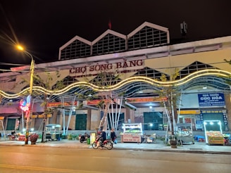 A lively street market scene at night featuring a building with the sign 'CHỢ SÔNG BẰNG'. There are various food stalls lit with bright lights, a few parked motorcycles, and people engaging in activities. Neon lights and trees are visible along the sidewalk.