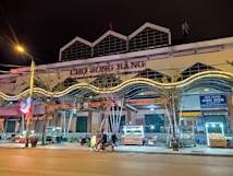 A lively street market scene at night featuring a building with the sign 'CHỢ S&Ocirc;NG BẰNG'. There are various food stalls lit with bright lights, a few parked motorcycles, and people engaging in activities. Neon lights and trees are visible along the sidewalk.