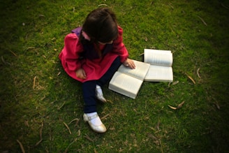A child wearing a bright red dress sits on green grass, surrounded by open books. The child appears to be reading or examining one of the books. The scene is serene and suggests a peaceful moment of learning or exploration.