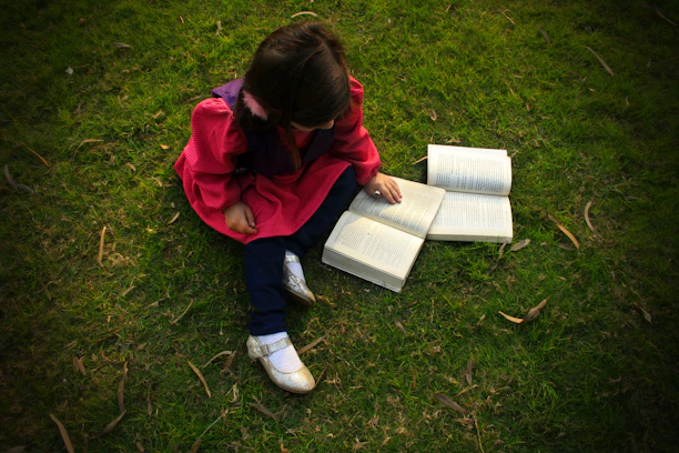 A joyful child sitting cross-legged, reading a bright, illustrated storybook under a tree.