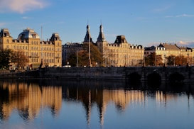 A serene cityscape features classic European architecture with buildings adorned with spires and intricate designs. The structures are beautifully reflected in a calm body of water in the foreground. Autumn trees add warm hues, and the scene is bathed in the golden light of the setting sun.