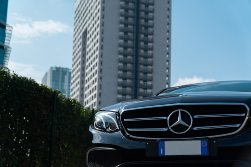 A luxury car with a prominent grille and emblem is parked in an urban environment. Tall modern buildings rise in the background against a partly cloudy sky. There appears to be a hedge or greenery on one side.