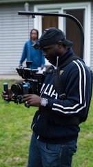 A person wearing a cap and a tracksuit is operating a professional camera rig outdoors. The setup includes a camera stabilizer system, and the person appears focused on filming. In the background, another person stands near a building with wood siding and a glass door.