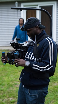 A person wearing a cap and a tracksuit is operating a professional camera rig outdoors. The setup includes a camera stabilizer system, and the person appears focused on filming. In the background, another person stands near a building with wood siding and a glass door.
