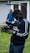 A person wearing a cap and a tracksuit is operating a professional camera rig outdoors. The setup includes a camera stabilizer system, and the person appears focused on filming. In the background, another person stands near a building with wood siding and a glass door.