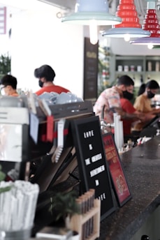 A cozy cafe environment with pendant lights hanging from the ceiling. The counter has various items, including a letter board with a message, cups, and writing utensils. People wearing masks are engaged in activities behind the counter, possibly preparing or serving food and drinks.