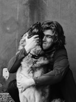 A person with medium-length hair embraces a large dog, holding its face affectionately. The image is in black and white, highlighting the textures and the bond between them.