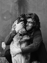 A person with medium-length hair embraces a large dog, holding its face affectionately. The image is in black and white, highlighting the textures and the bond between them.