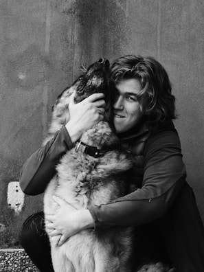 A person with medium-length hair embraces a large dog, holding its face affectionately. The image is in black and white, highlighting the textures and the bond between them.