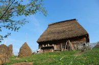 A rustic wooden house with a thatched roof surrounded by wooden fencing and haystacks. The landscape is green with patches of yellow flowers, and a clear blue sky is above. A tree with green leaves is visible on the left side.