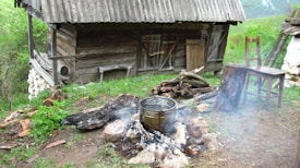 A rustic wooden cabin with aged wood and a slanted roof stands in a grassy area. In front, a makeshift outdoor fireplace visible with a metal pot heating over smoldering logs surrounded by stones. To the side, simple wooden furniture including a chair and a table made from rough wood, adding to the rustic atmosphere. The scene is set in lush greenery, with plants and forested hills in the background, suggesting a remote, natural setting.