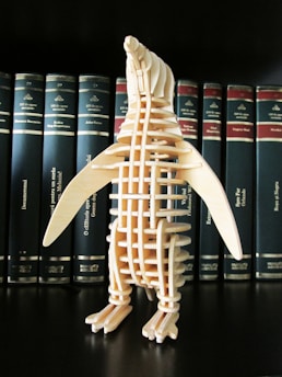 A wooden penguin model stands in front of a row of hardcover books with dark blue spines and gold text. The model is composed of several interlocking wooden pieces, giving it a skeletal appearance.