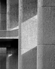 A close-up view of a textured concrete architectural structure, featuring large columns and a geometrical play of light and shadows across the surface.
