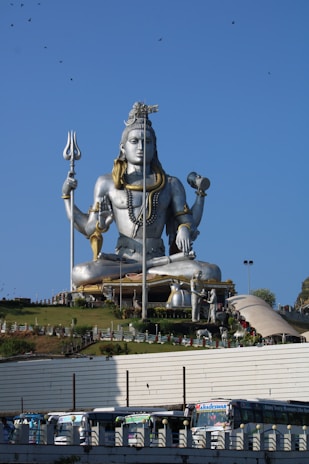 A massive statue of a deity, likely Shiva, is seated in a meditative pose atop a hill. The figure is adorned with gold ornaments and holds a trident. The sky is clear with a few birds flying, and there are people and trees visible in the foreground. Buses are parked at the bottom of the hill.