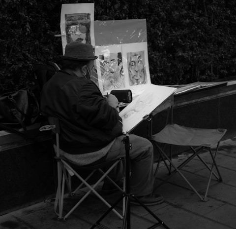 A person is seated on a folding chair, drawing or painting on a large sketchpad. Behind them are several portraits displayed on an easel, which appear to be sketches or paintings. The setting seems to be outdoors along a sidewalk or promenade, with a dense hedge in the background. The overall scene conveys a quiet, focused artistic environment.