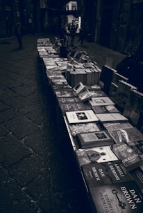 A narrow, cobbled street features a long table filled with a variety of books arranged in neat stacks. The setting appears to be a dimly lit alley with a few people walking in the distance; the atmosphere is moody and somewhat mysterious.