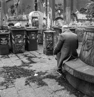 An elderly person sits alone on a stone bench in an urban setting surrounded by graffiti-covered walls and trash bins. The atmosphere is gritty and desolate, with a mix of modern urban decay elements like a payphone and recyclables processing bins marked with Italian labels.