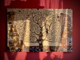 A painting featuring an intricate tree with swirling branches set against a rich, red backdrop. The artwork includes various geometric patterns, such as triangles and rectangles, along with decorative motifs. Shadow play on the canvas suggests sunlight streaming in, adding depth and dimension.