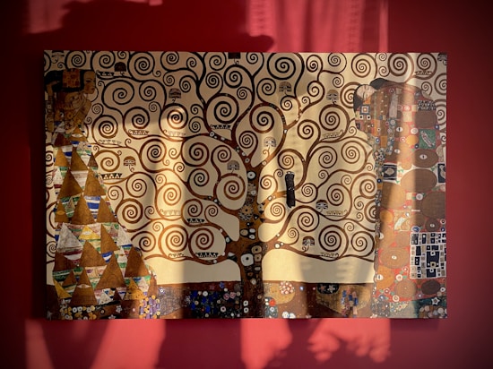 A painting featuring an intricate tree with swirling branches set against a rich, red backdrop. The artwork includes various geometric patterns, such as triangles and rectangles, along with decorative motifs. Shadow play on the canvas suggests sunlight streaming in, adding depth and dimension.