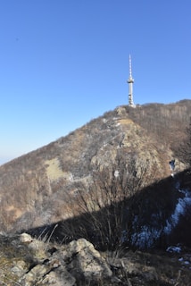 A telecommunications tower stands atop a rocky hill with sparse vegetation. The hill is mostly covered in dry foliage, with patches of snow visible. The sky is clear and bright blue, emphasizing the height of the tower.
