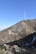 A telecommunications tower stands atop a rocky hill with sparse vegetation. The hill is mostly covered in dry foliage, with patches of snow visible. The sky is clear and bright blue, emphasizing the height of the tower.