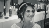 A woman with styled hair and a jeweled headband smiles broadly. She is wearing a detailed, beaded garment. The background displays various hairstyling products and equipment.