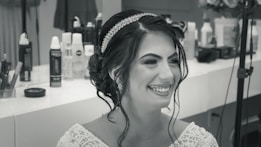 A woman with styled hair and a jeweled headband smiles broadly. She is wearing a detailed, beaded garment. The background displays various hairstyling products and equipment.