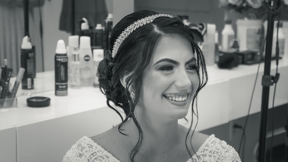 A woman with styled hair and a jeweled headband smiles broadly. She is wearing a detailed, beaded garment. The background displays various hairstyling products and equipment.