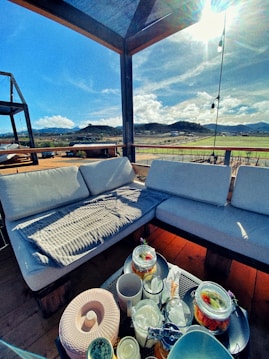 A cozy outdoor seating area is set beneath a shaded pergola, surrounded by a scenic countryside landscape. The sky is bright and sunny, casting natural light over the wooden deck and white cushioned sofas. On the table, there are various jars containing fruit and possibly infused drinks, with cups and a large sun hat placed beside them.